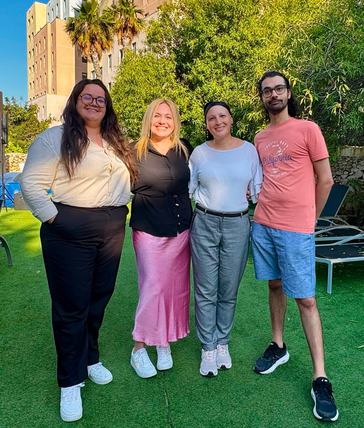 Four people smiling together on a sunny garden lawn with palm trees during the EPIC-X mobility visit in Malta